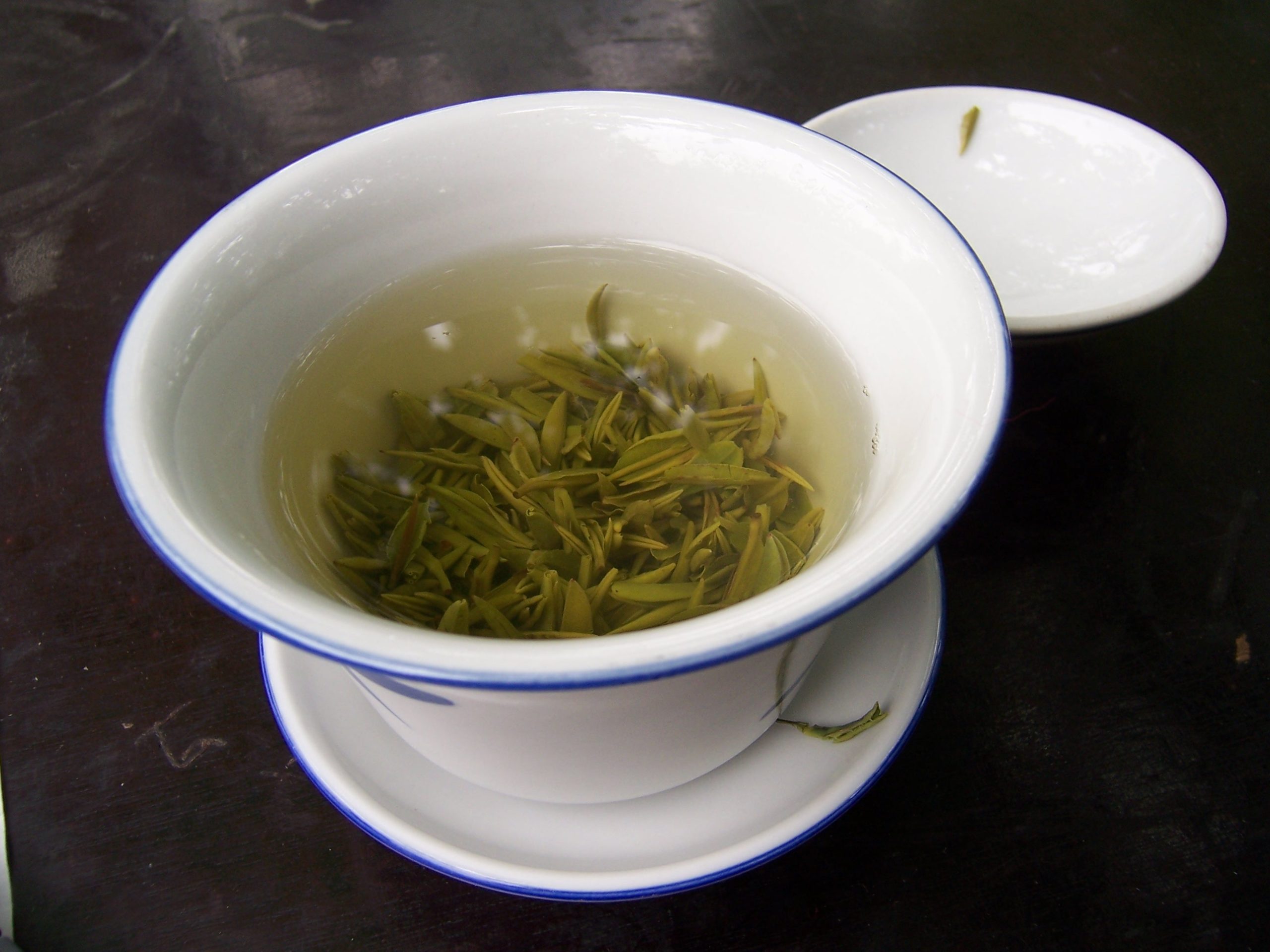 Green tea leaves unfurling in hot water in a white lidded cup with a blue rim