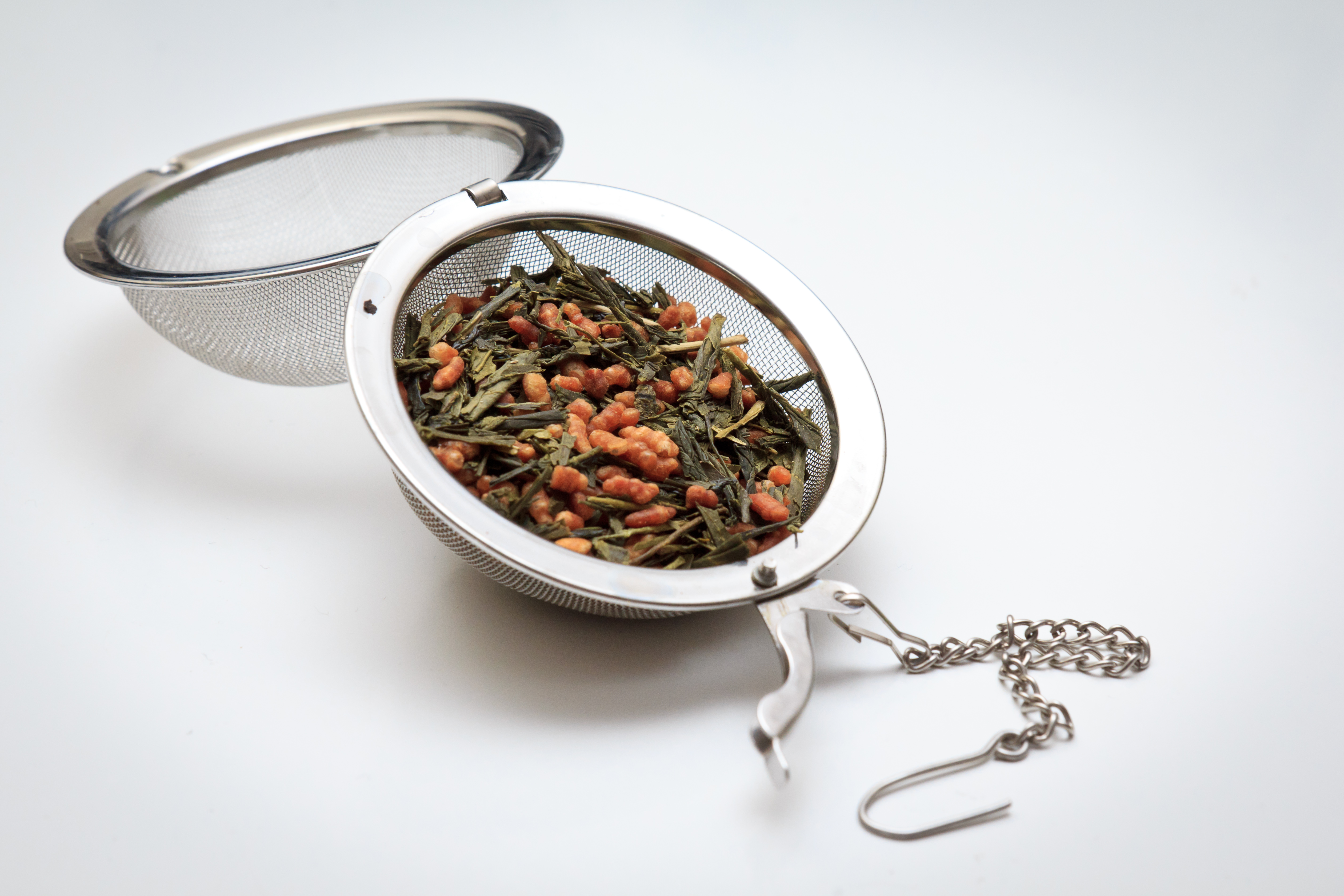 An open mesh tea infuser ball filled with green tea leaves and toasted rice