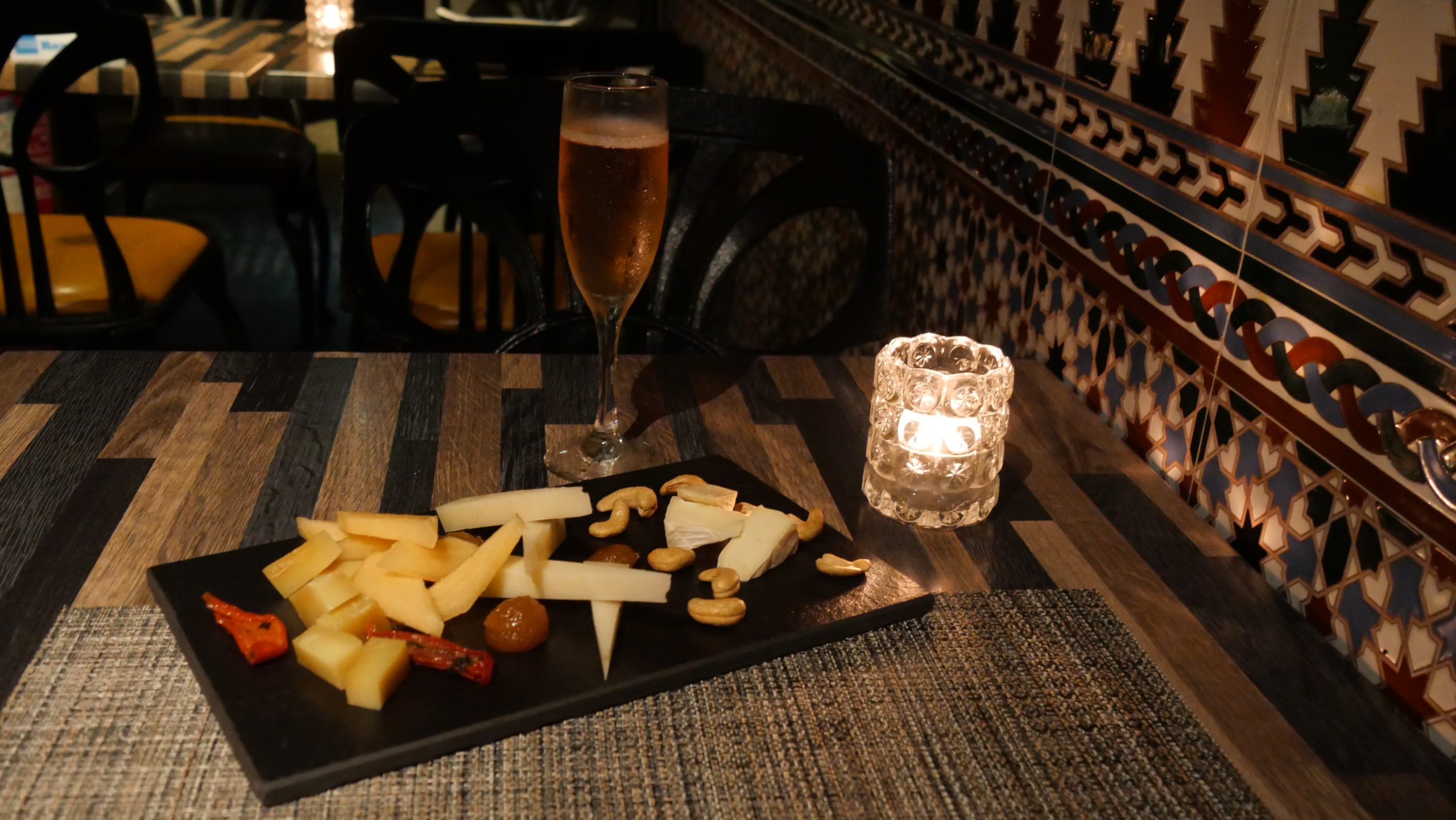 A slate of sliced cheeses and cashews beside a flute of sparkling rosé and a candle on a restaurant table