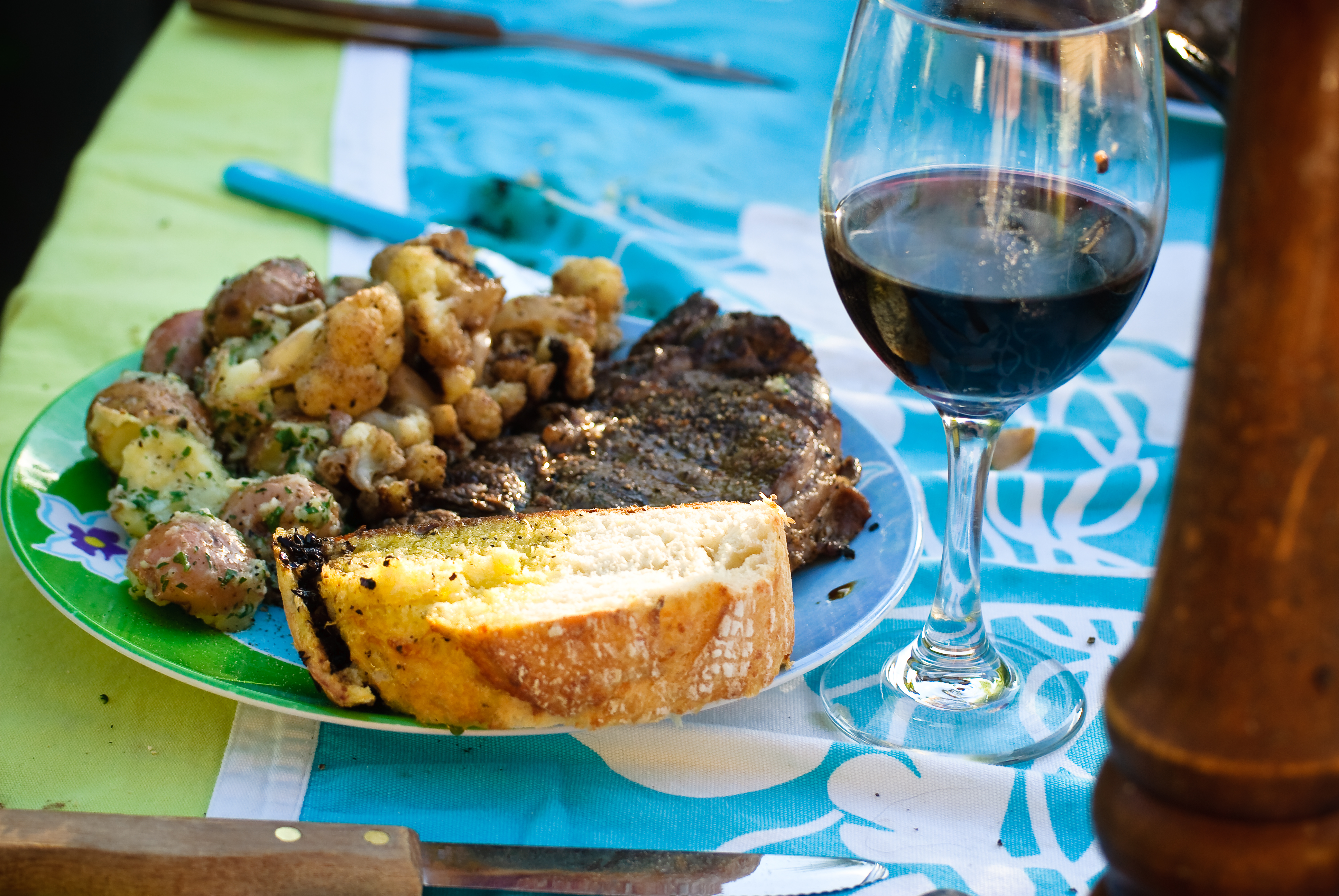 A grilled ribeye steak with roast potatoes, cauliflower and garlic bread beside a glass of red wine