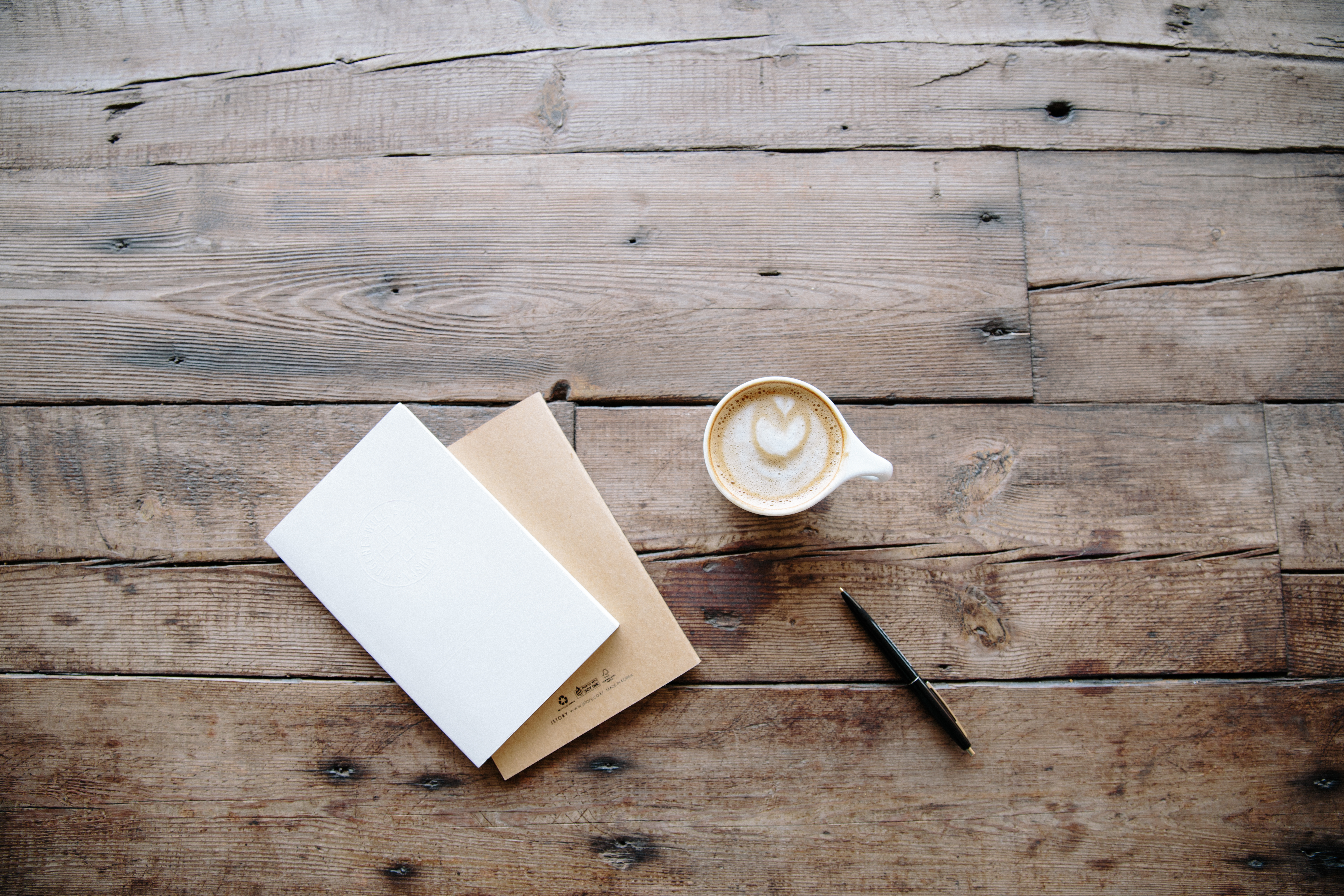 A cup of coffee with latte art, two notebooks and a pen on a weathered wooden table seen from above