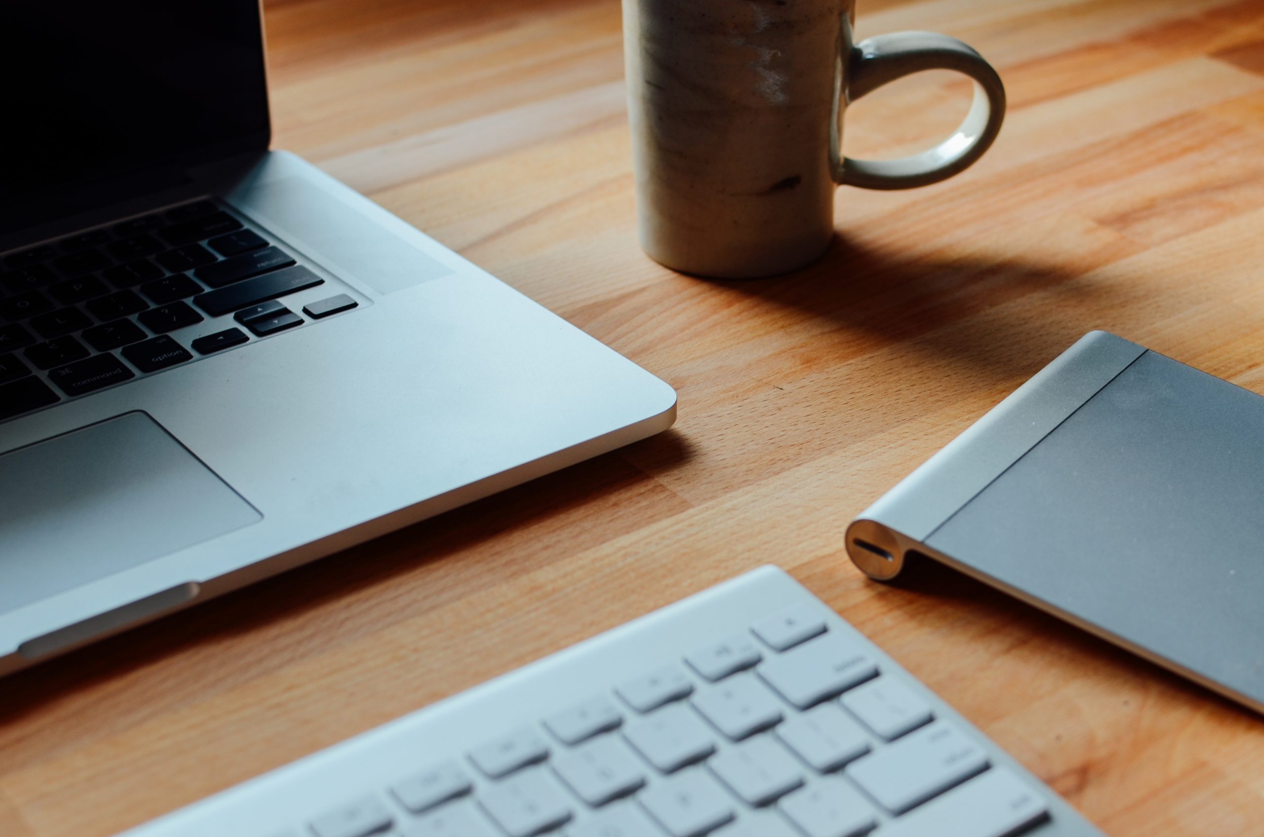 An open laptop, a stoneware mug, a wireless keyboard and a trackpad on a warm wooden table