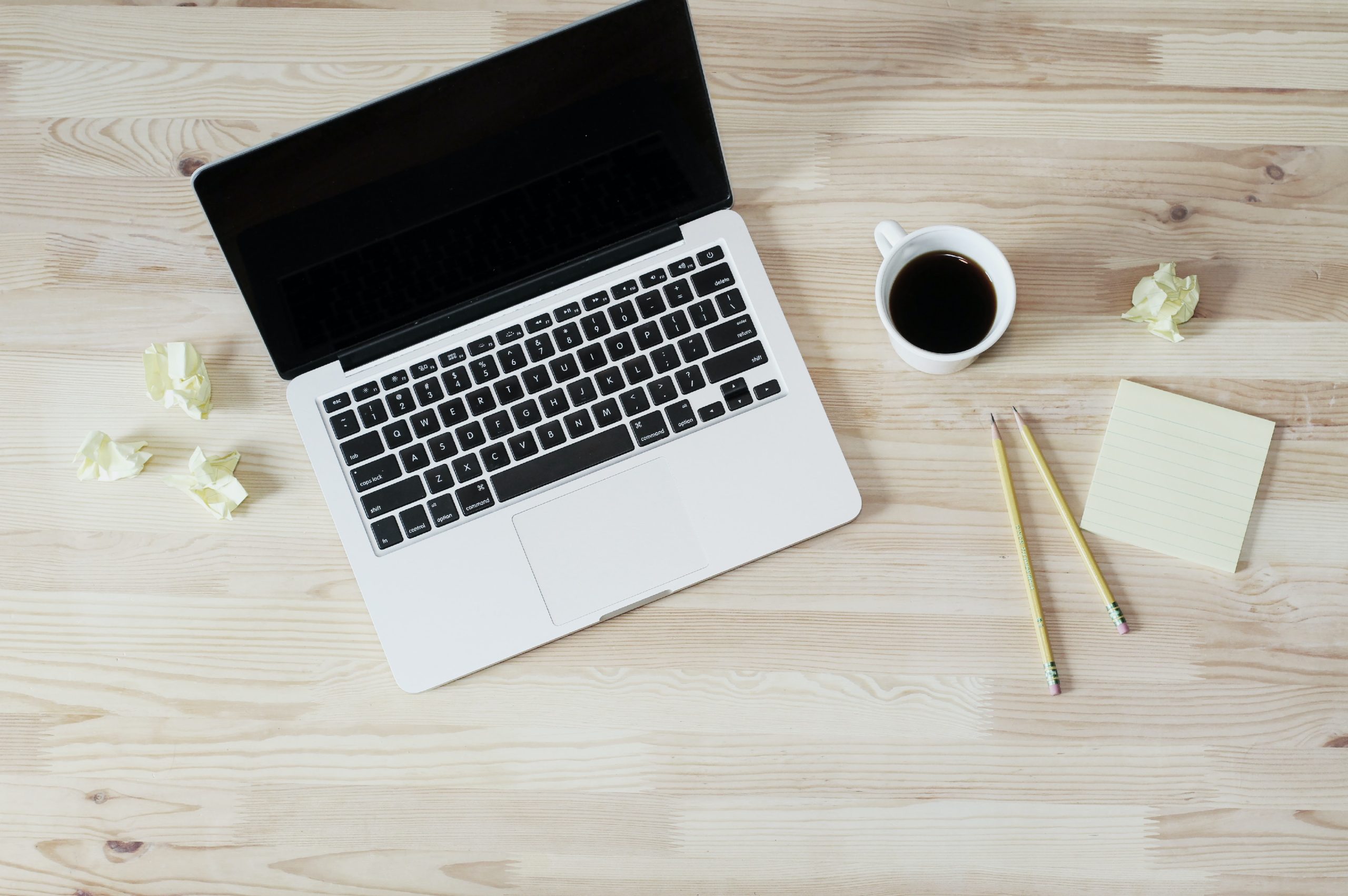 Overhead view of a laptop, a cup of black coffee, two pencils, a sticky note pad and crumpled paper on a wooden desk