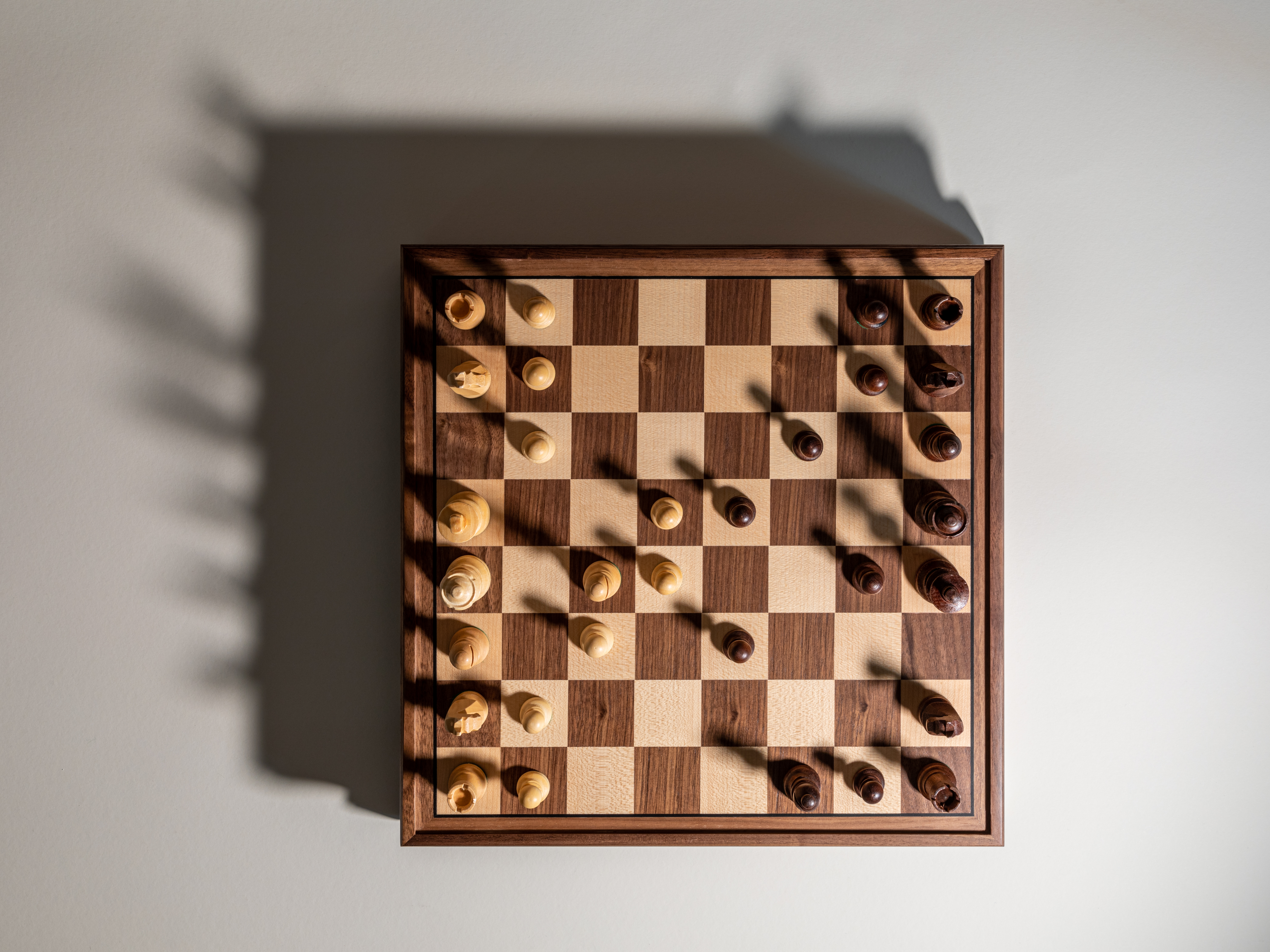 Overhead view of a wooden chessboard mid-game casting a long shadow across a pale table