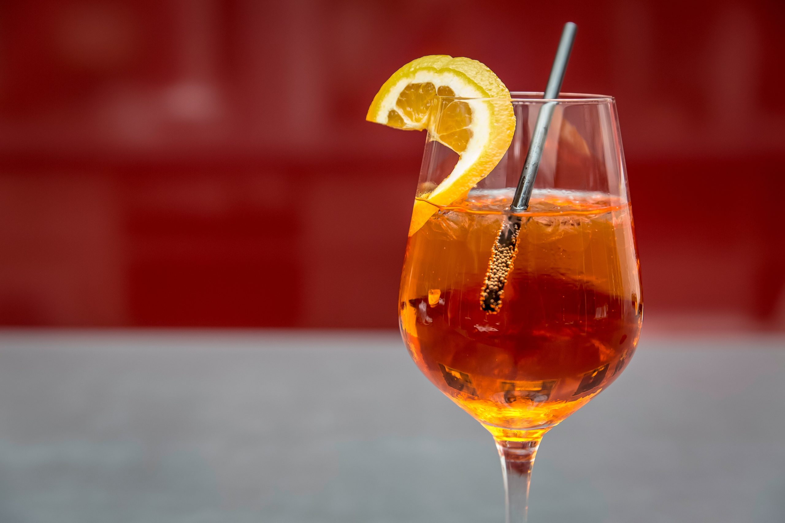 A stemmed glass of amber iced tea with ice, a slice of orange and a metal straw against a red background