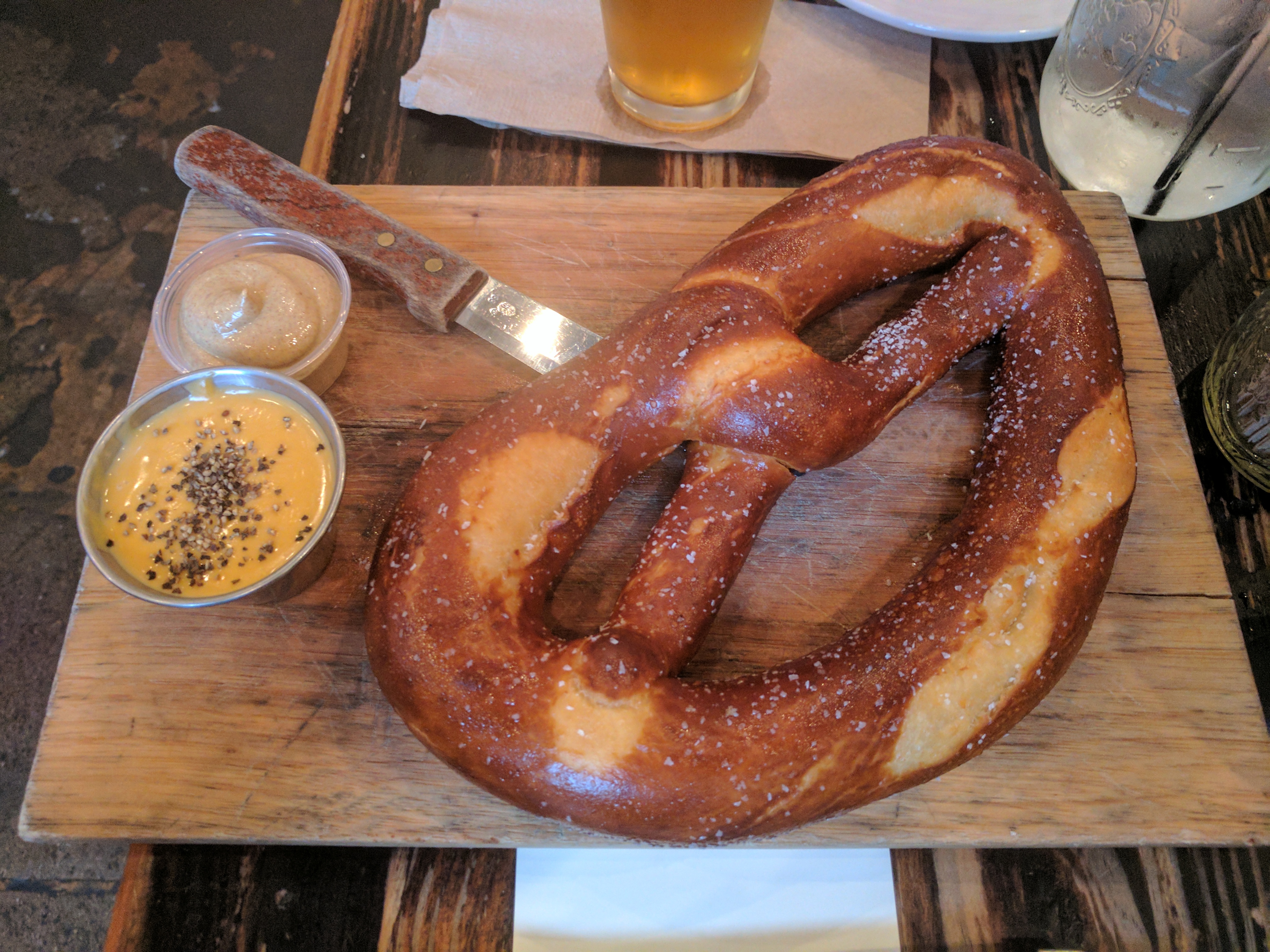 A large salted pretzel on a wooden board with cheese sauce and mustard beside a glass of pale beer
