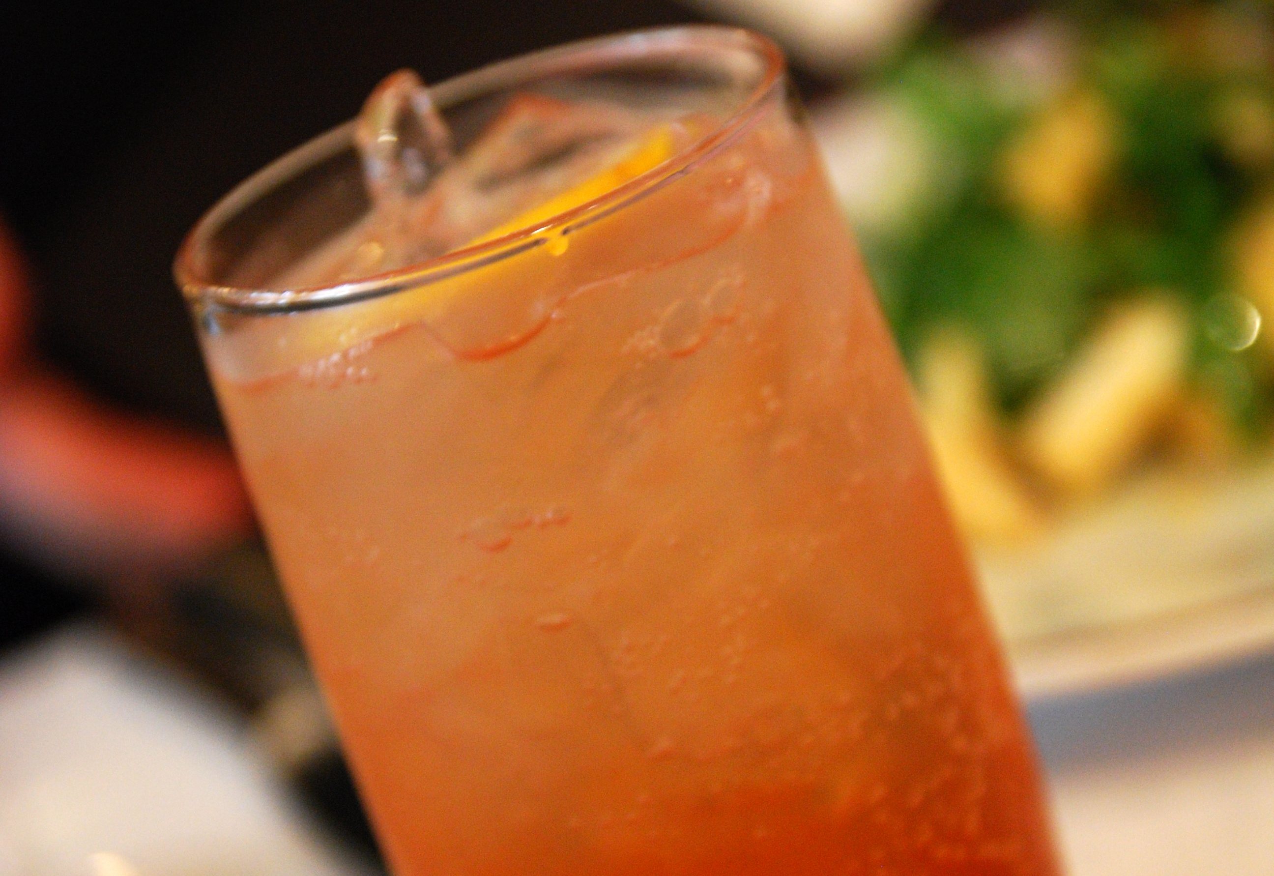 A tall glass of pale pink sparkling drink with ice and a slice of lemon beside a plate of food