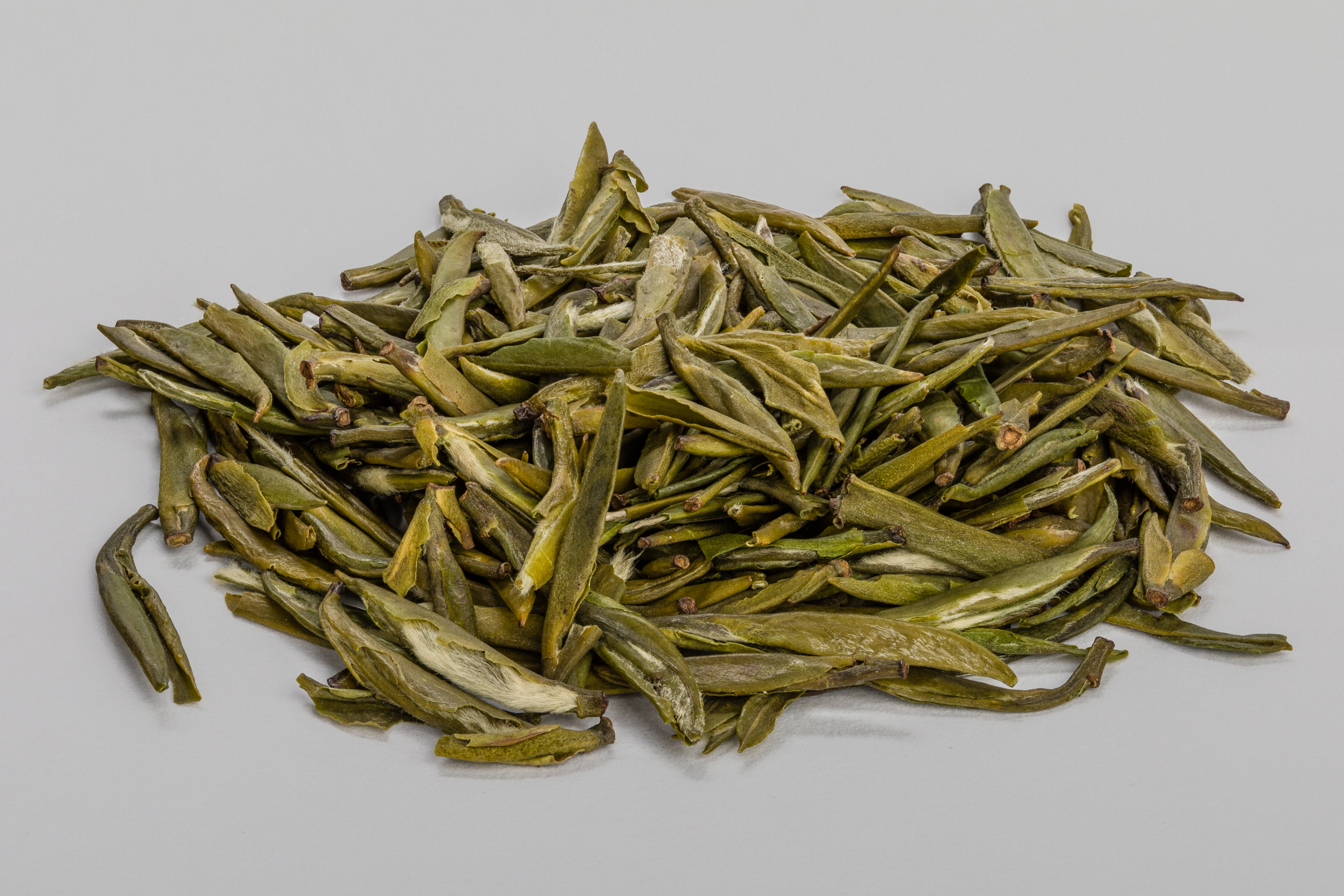 A small heap of long, flat green tea leaves on a pale gray surface