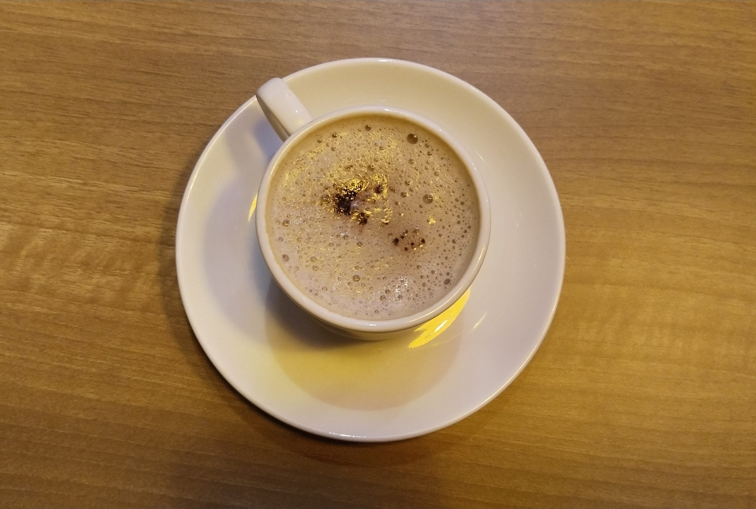 Overhead view of a white cup of frothy milky coffee on a saucer on a light wooden table