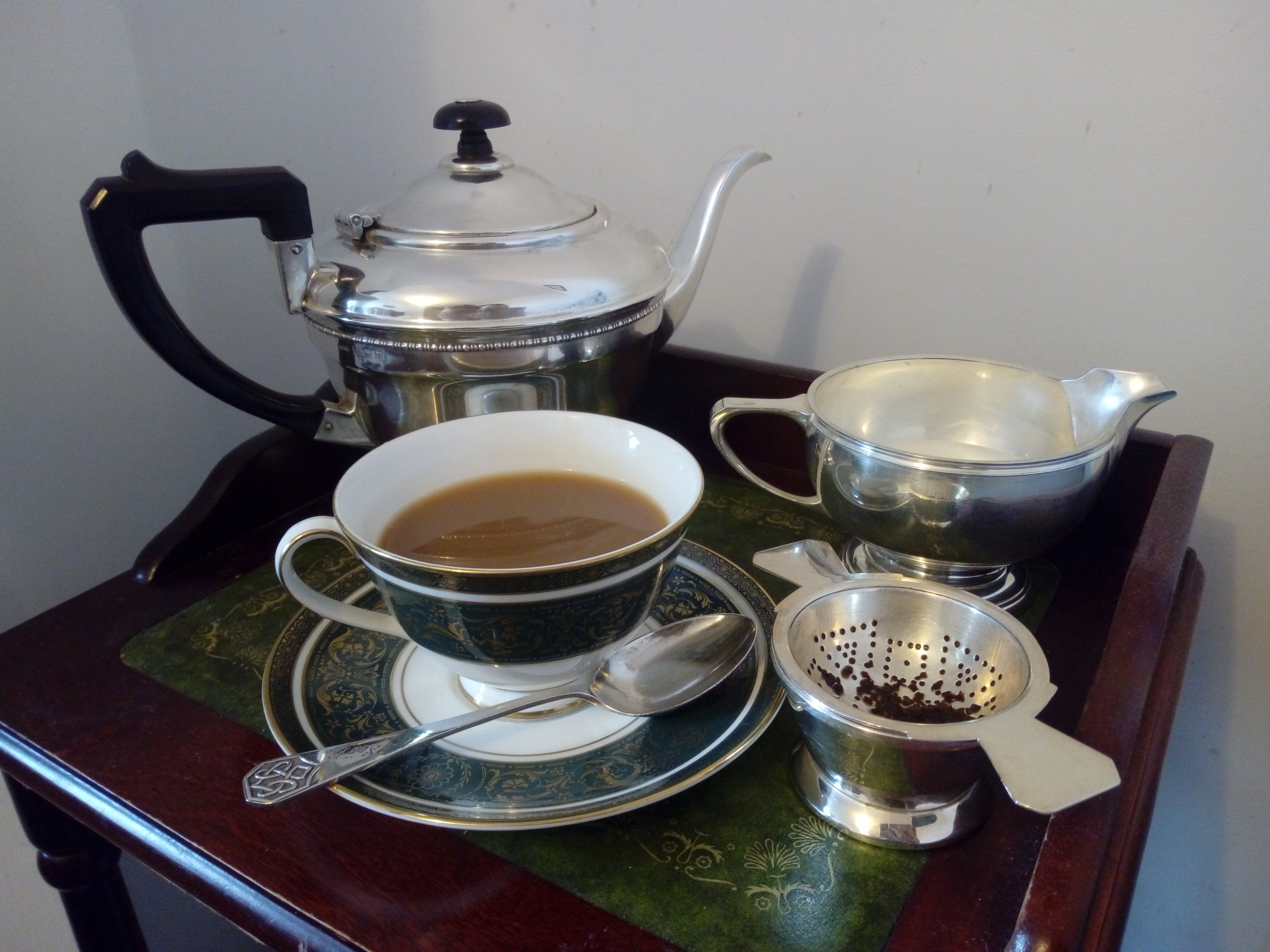 A silver teapot, milk jug and tea strainer beside a patterned cup of milky tea on a saucer on a wooden tray