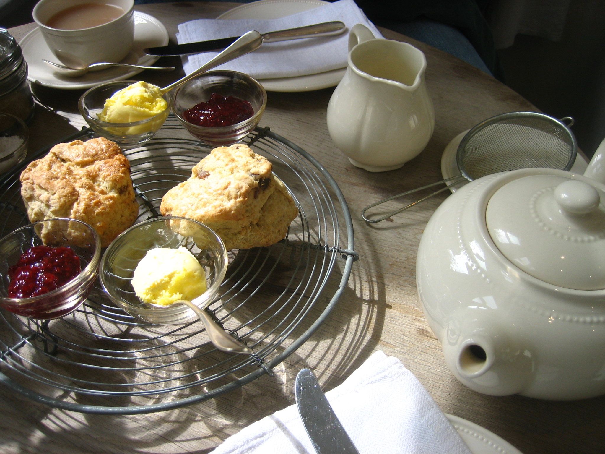 Scones on a wire rack with butter and jam beside a cream teapot, milk jug and tea strainer