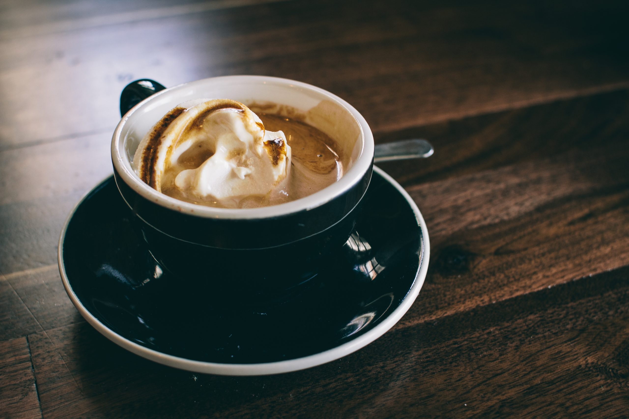 A dark cup of coffee topped with a scoop of toasted cream on a black saucer on a wooden table