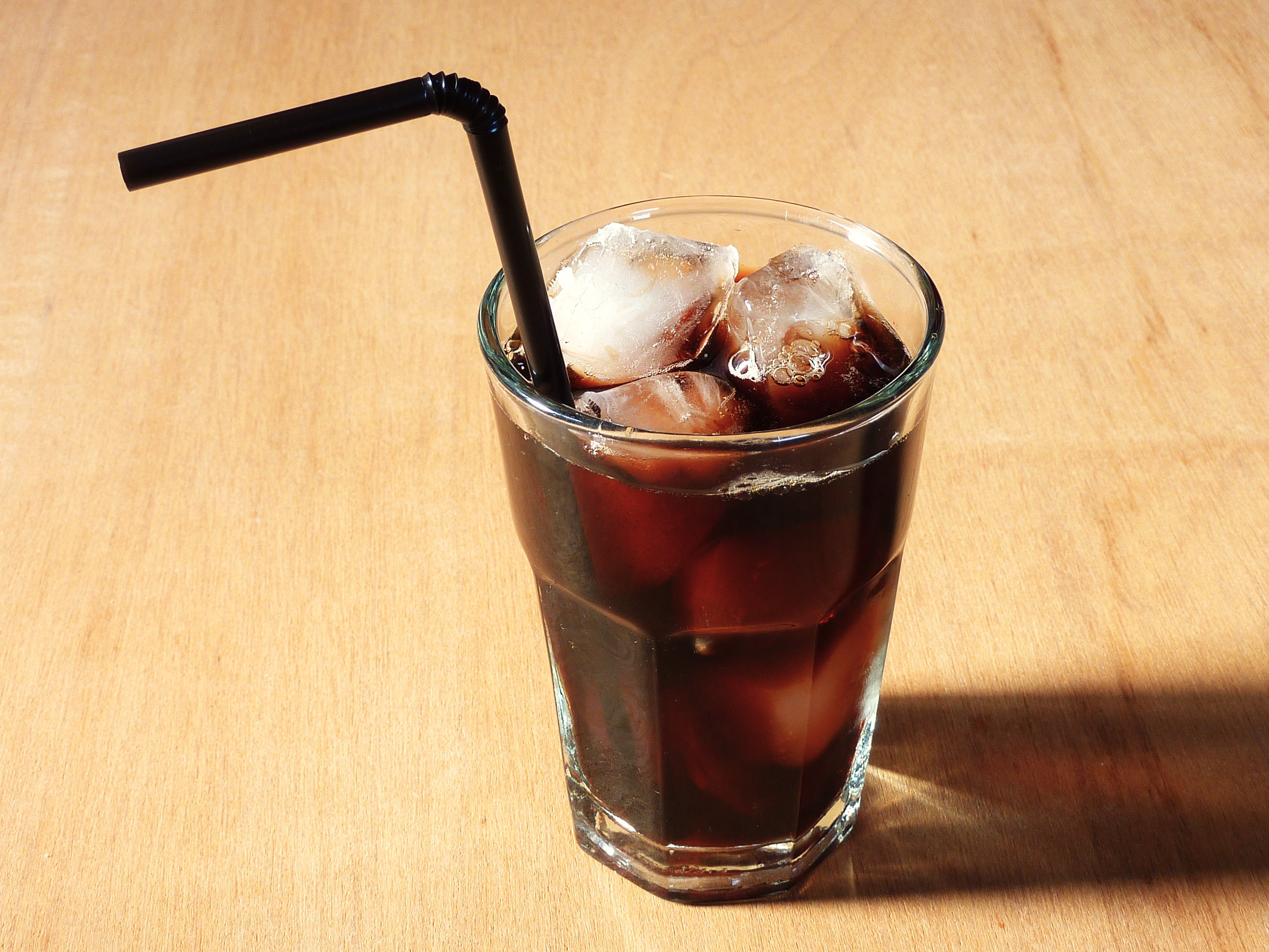A glass of black iced coffee with ice cubes and a black straw on a light wooden table