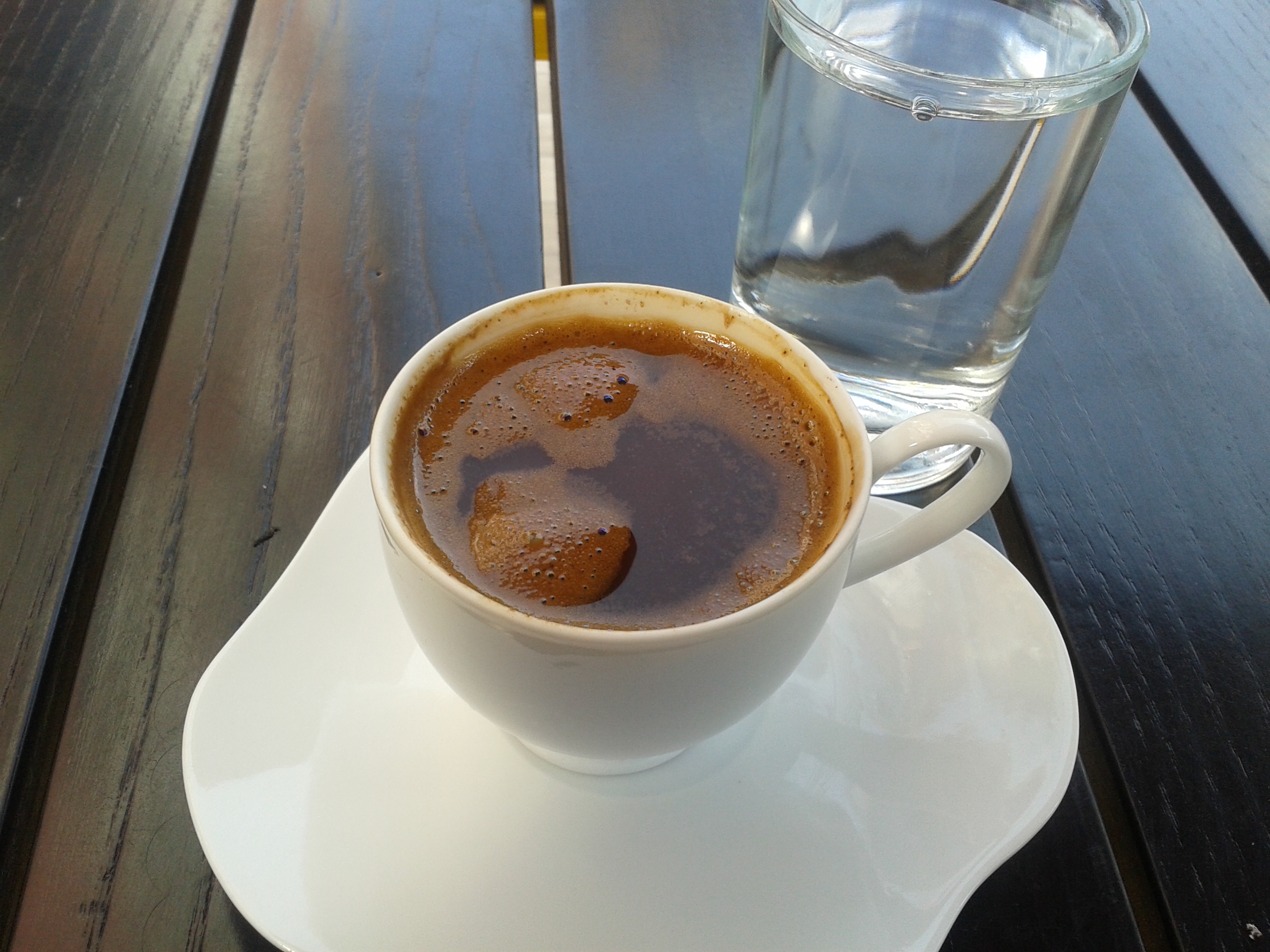 A small white cup of Turkish coffee on a saucer beside a glass of water on a dark wooden table