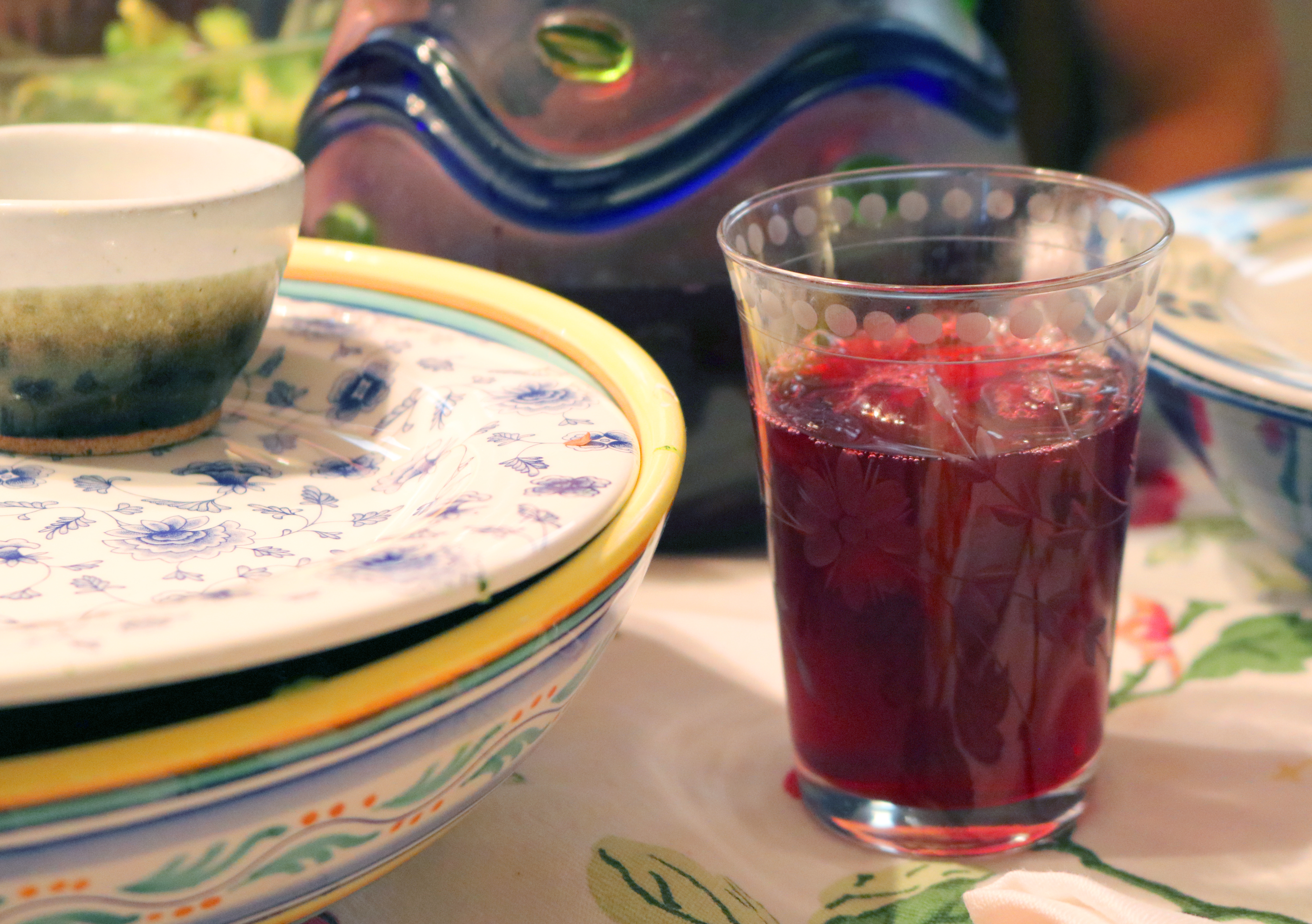 A glass of deep red hibiscus tea with ice on a table beside patterned plates