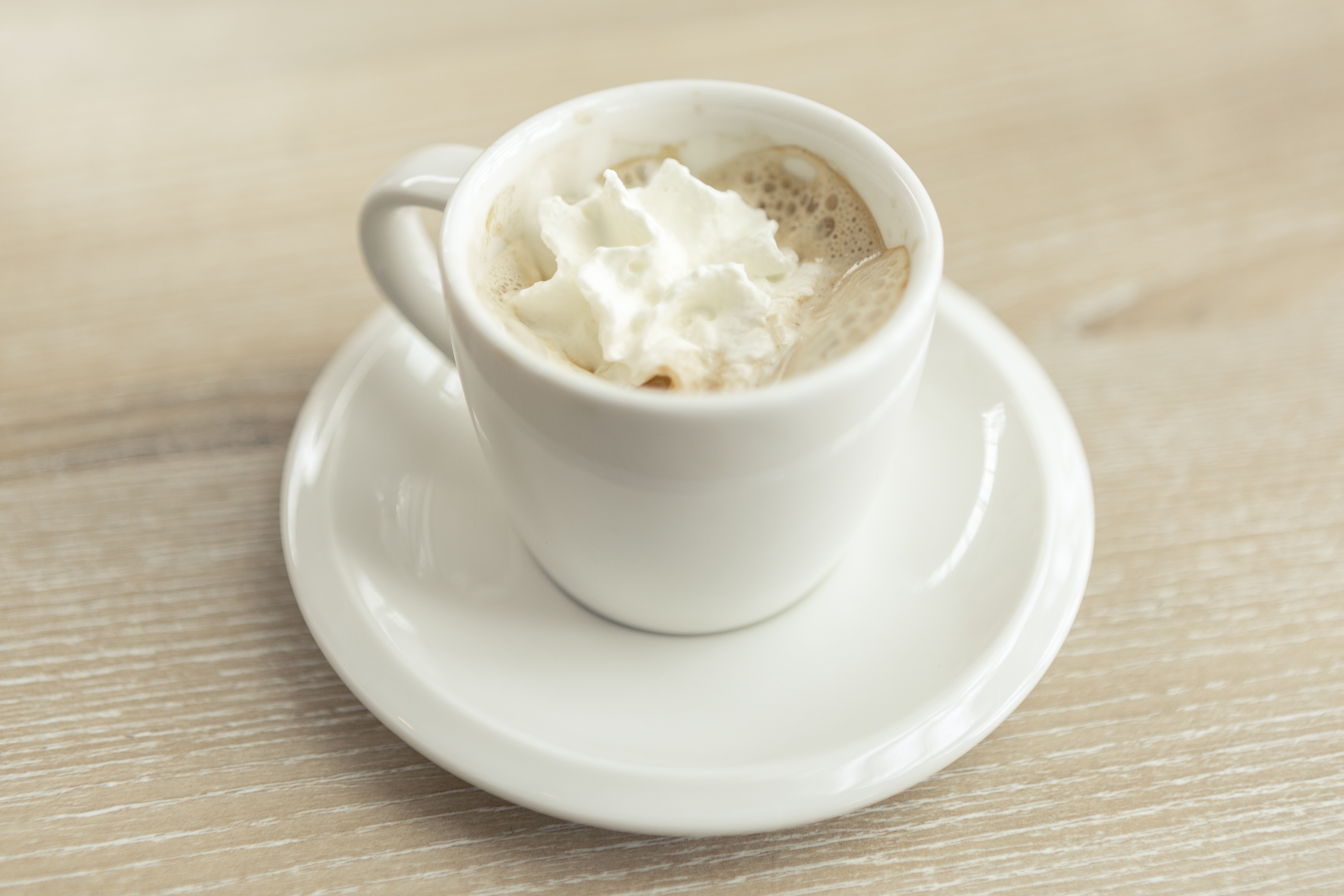 A small white cup of espresso topped with whipped cream on a saucer on a pale wooden table