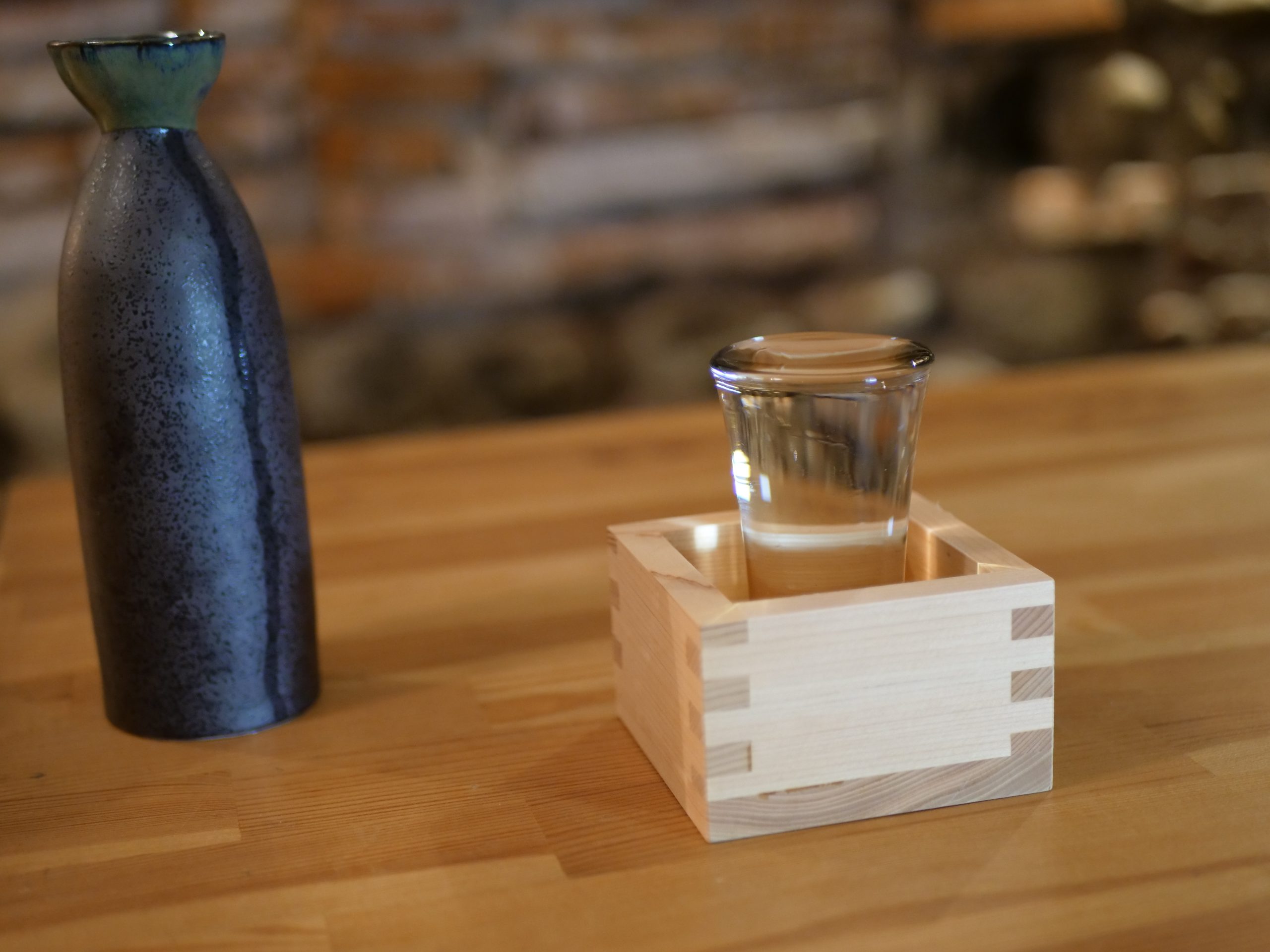 A dark ceramic sake flask beside a small glass of clear sake set in a wooden masu box