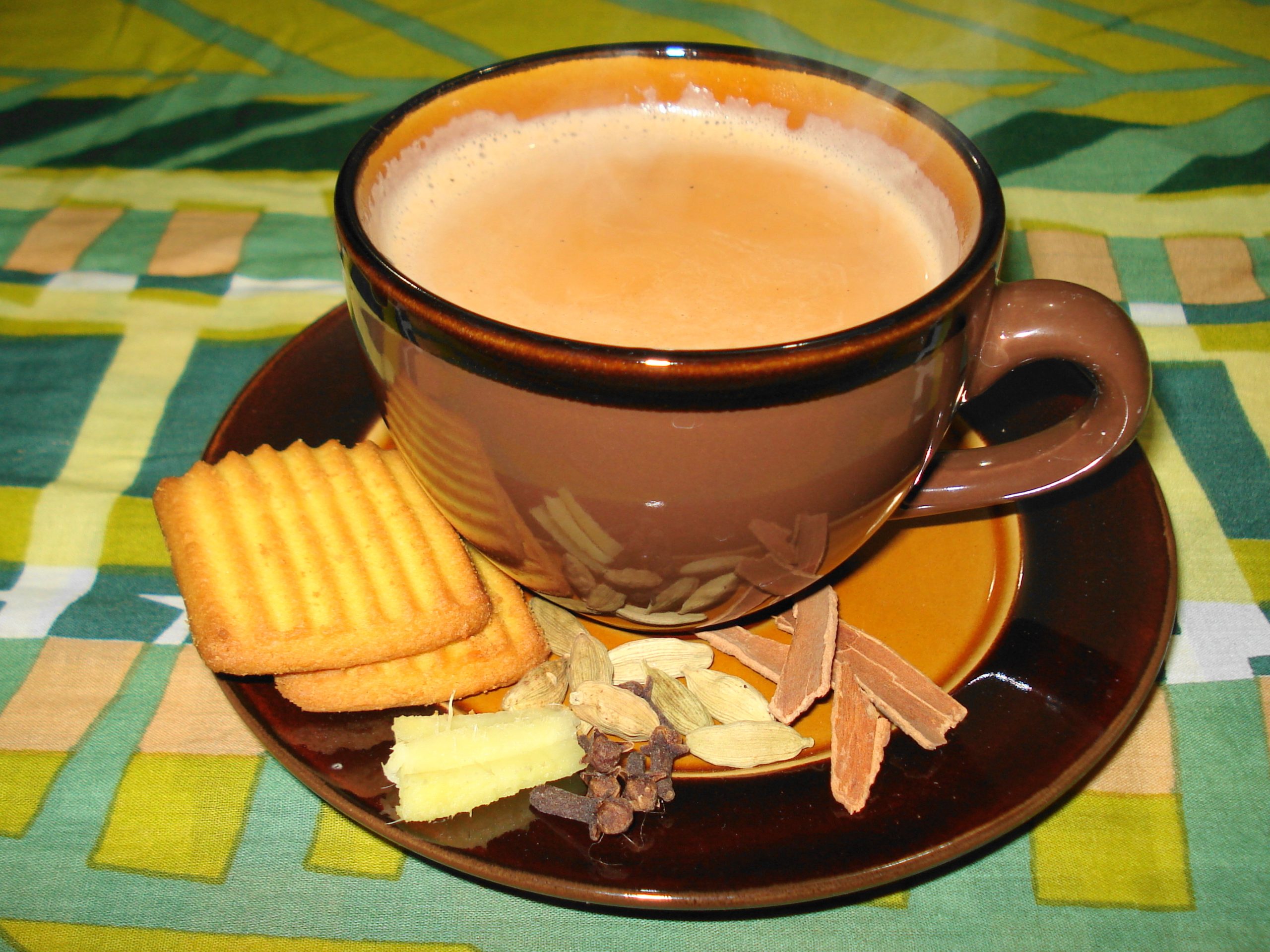A cup of milky masala chai on a saucer with biscuits, cardamom pods, cloves, cinnamon and fresh ginger