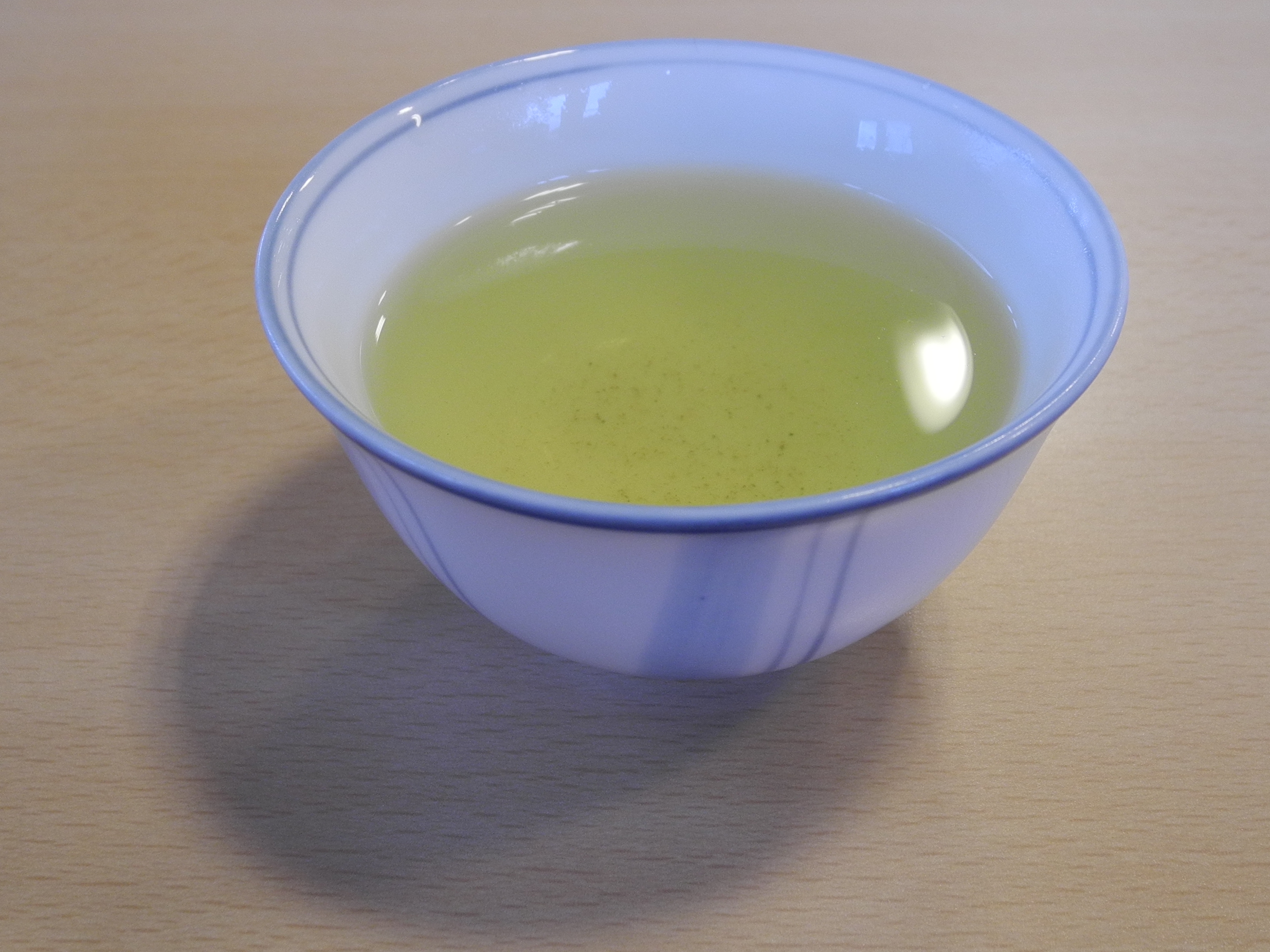 A small white bowl-shaped cup of pale green tea on a light wooden table